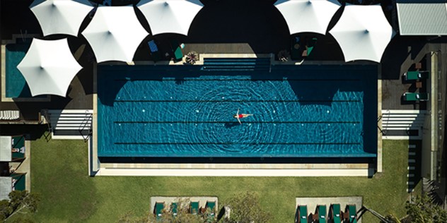 An aerial view of a pool with dark blue water, surrounded by grass and and six white umbrella tops. A woman in a one-piece red bathing suit floats in the middle of the pool. 