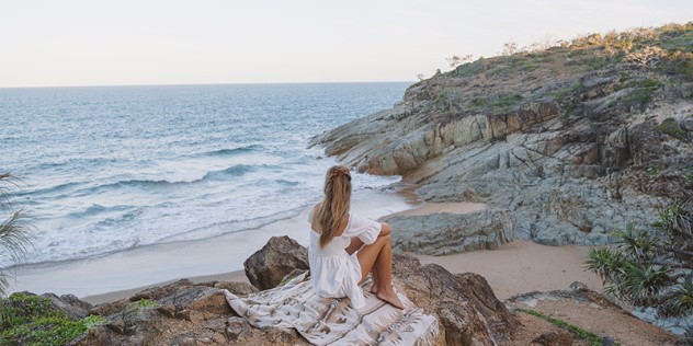 A blond woman in a white dress sits on a patterned rug on rocks along a small beach on a clear day, looking out at the water.