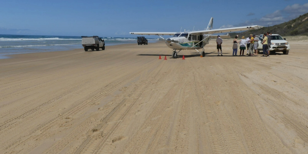  People standing next to a biplane parked on a wide, sandy beach with raised, grassy shores.