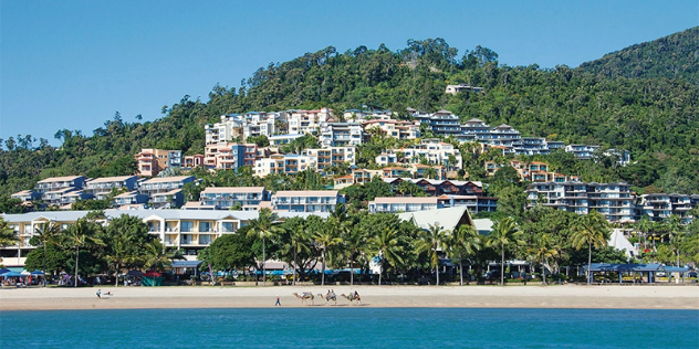  A tree-covered hill with apartments and hotels built into it, leading to a white sand beach. 