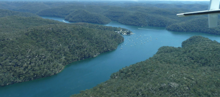 An aerial view of a bay splitting into two estuaries, that forms a t-shape of deep blue water surrounded by green treed hills, on a clear day.