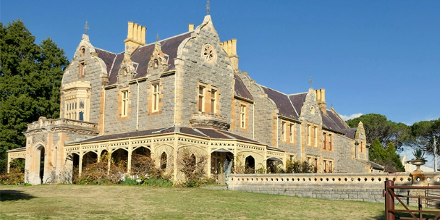  A heritage-listed large, stone Victorian house, with a large veranda, framed by grass, trees and blue sky.