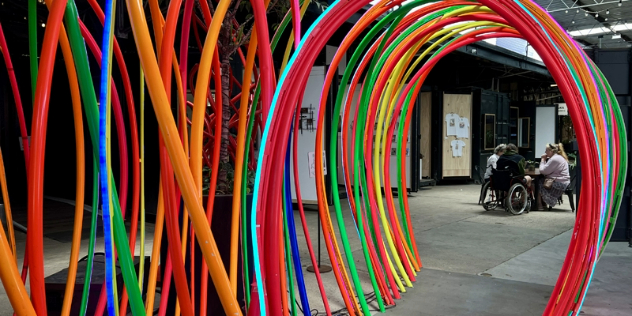  A wheelchair user and two others sit at a table as seen through a tunnel formed by colourful tubes curbed and stuck to the floor. 