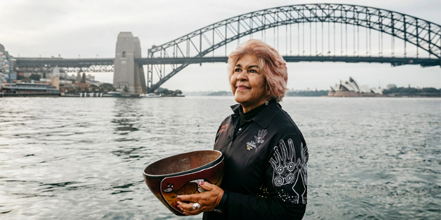 Aunty Margaret, holding a bowl decorated with Aboriginal designs, standing along the shore with the Sydney Harbour Bridge behind her. 