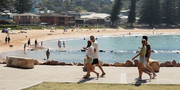 Two couples walk along a concrete boardwalk on a sunny day in front of a sandy, curved beach dotted with umbrellas and people, with homes and trees in the background.