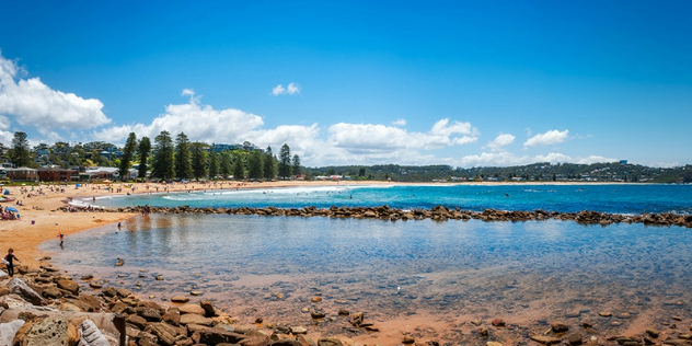  A sunny, tree lined beach dotted with people, on a curved bay divided by a line of rocks. 