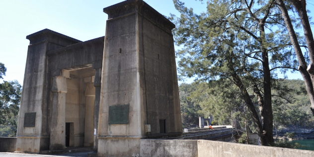  A concrete gate created by two pillars over the entry to a bridge over a dam, framed by trees and blue sky. 