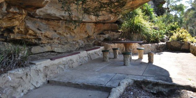  A round picnic table and chairs moulded from rocks to match the rock wall next to it, surrounded by sun-lit foliage. 