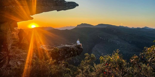 Sunset seen over mountains through a rock formation shaped like the letter C jutting into the air over a valley, a woman sits precariously on the bottom edge.