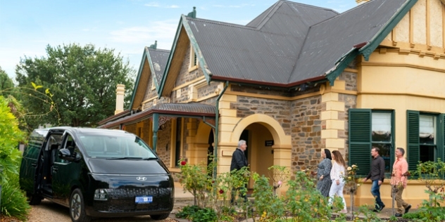 black SUV parked in the driveway and a group of people walking towards an 1890s victorian sandstone building with tin roof