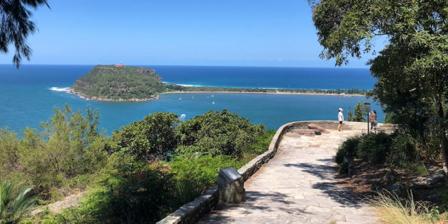 A stone path flanked by trees leads to a lookout over the ocean, where a single island-like tree-covered mound can be seen, attached to shore by a skinny peninsula of land.