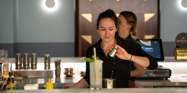 A white bartop with a single tall-glass cocktail on it, while a female bartender is in the process of tossing a shot of liqueur into a tumbler in the background behind the bar.