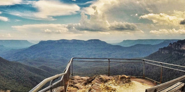  A lookout point with a chain metal fence juts out over a canyon of treed valleys and flat topped mountains.