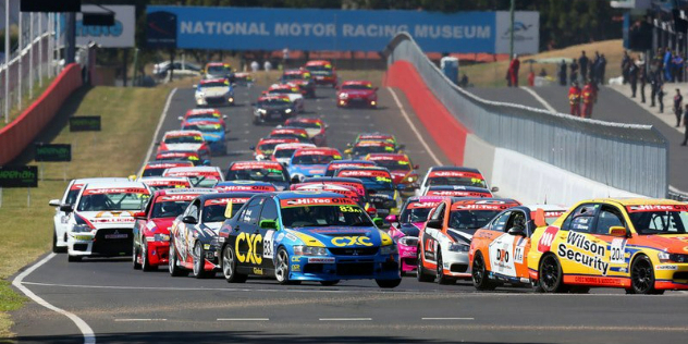  Dozens of colourful racecars rounding a bend along a track, with pit crews looking on.