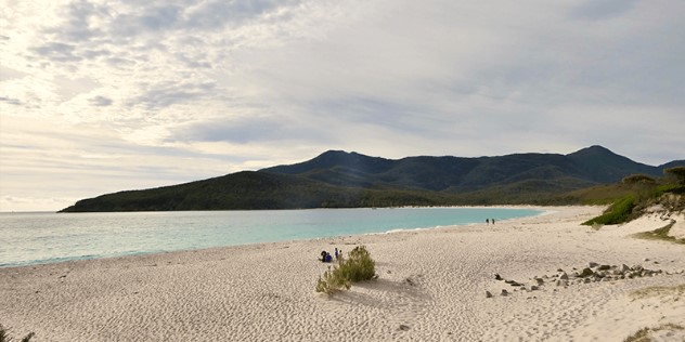 A turquoise blue bay of water sweeping up to white sand beach with bits of green scrub, with two points of green, treed mountains rising in up on the other side of the bay. 