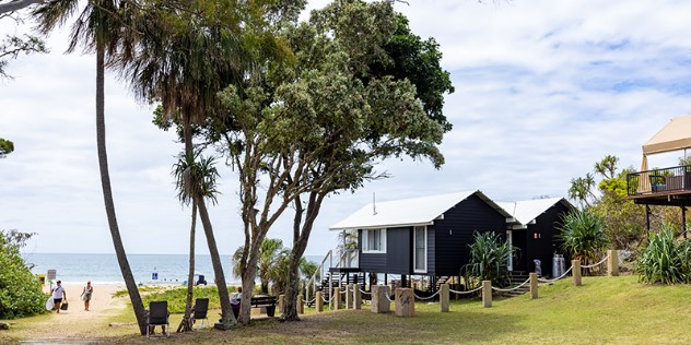 A dirt path opens to a beach and sea on the horizon, next to it are two beach houses on stilts, facing the water.