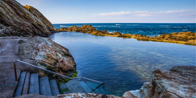 A man-made flat path along a rock wall leads to a metal guardrail and steps into a natural rock pool, with the ocean and blue sky on the horizon.