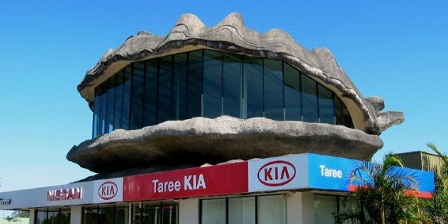 A thee metre tall, half open oyster sculpture, with the open mouth lined with tall dark windows, sitting on top of the Taree KIA dealership building, under blue sky.