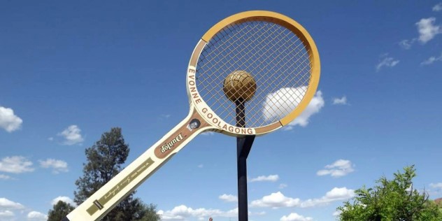 A fourteen metre long, white and tan trimmed tennis racquet mounted in the ground and tilted on its side as though it’s hitting the giant ball on a pole behind it.