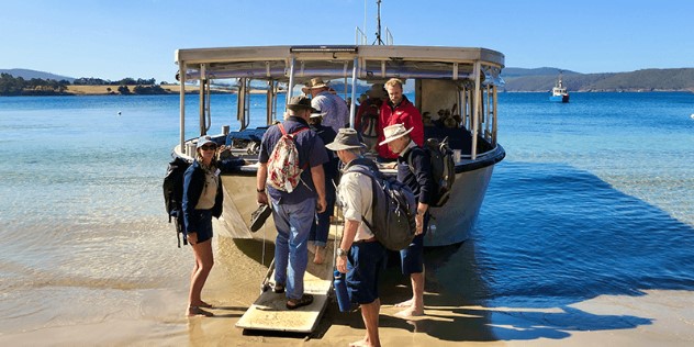 A small tour group of people in hats walking up a ramp to get on a smaller-wooden tour boat docked on a sandy beach under blue sky. 