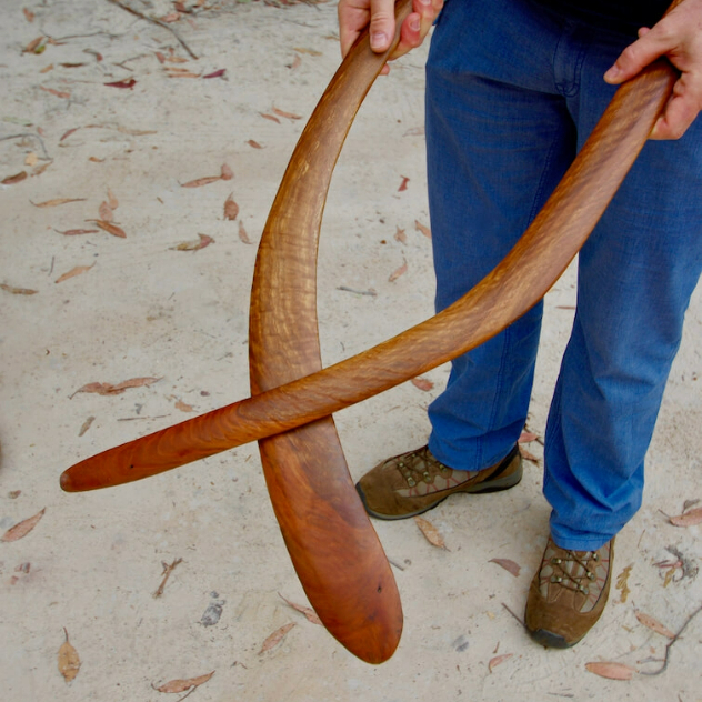  A man in jeans holding two long, handmade wooden boomerangs over a sandy beach.