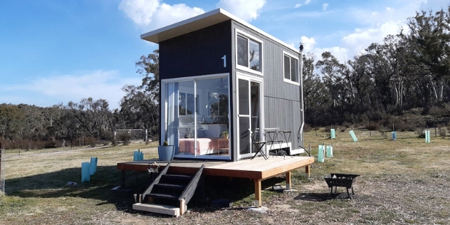 narrow, grey, weatherboard, two storey tiny home with large windows positioned to optimise the rural view