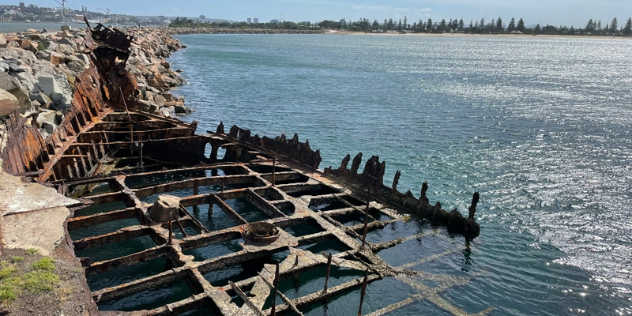  A rusted skeleton of the bottom of a ship, pressed up against a break wall of rocks dividing a sunny lake. 