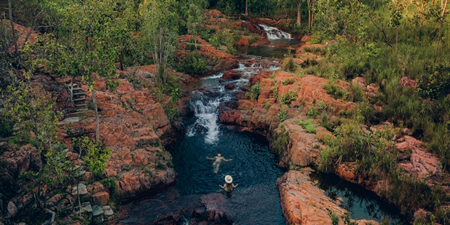 A small waterfall over a red-rocky creek with man-made steps along the edge, leading to a waterhole where a couple is swimming.