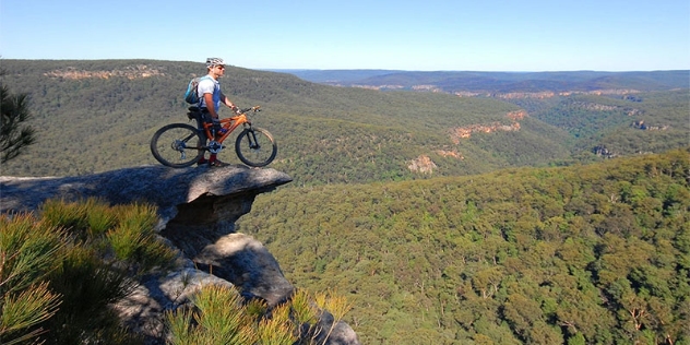 man with orange mountain bike looks down at tree tops from rocky lookout