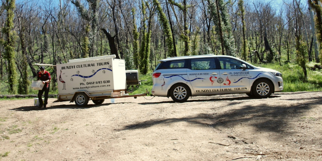  A man unloading a car and trailer on a sunny bush road, with Aboriginal designs and Bundyi Cultural Tours painted on both.