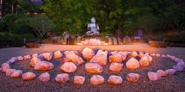 view of rock crystals placed in a circle pattern on gravel with illuminated crystals at the feet of a stone buddha in the gardens at dusk