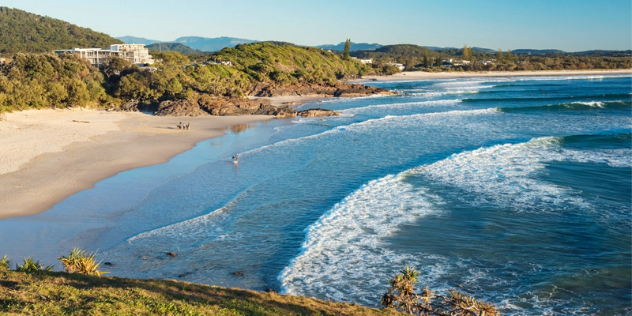 A few surfers on a sunny, sandy beach with rocky outcrops, a modern house is perched on green cliffs behind them.