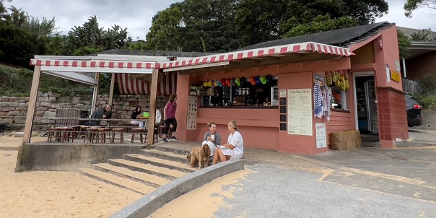 A terra-cotta coloured brick, beachside cafe and kiosk for travel information, with a small patio of tables and chairs visible in the back and in front a passthrough window and sandy-covered steps leading onto the beach. 