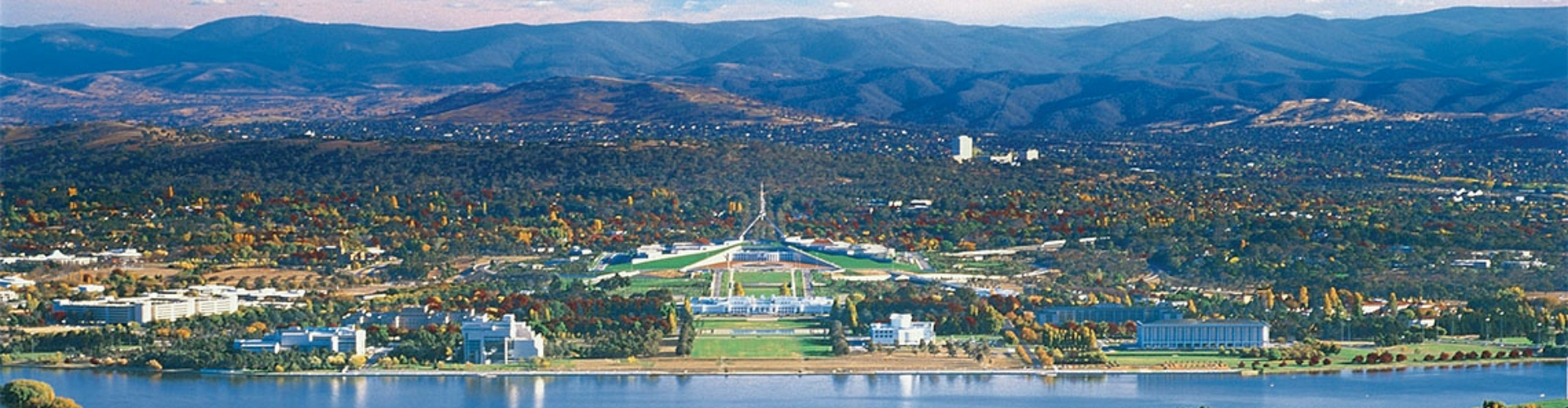canberra-parliament-and-city-act-1920x500 A panoramic view of Canberra city, with parliament house in the centre in front of a lake, with flat treed plains and a mountain range behind it.