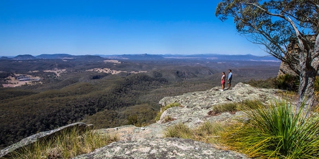 two people on a cliff top lookout over a valley of trees with distant hills