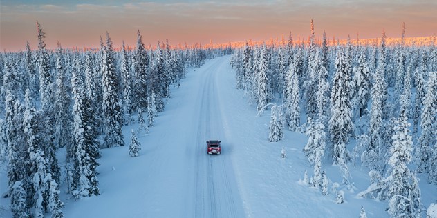 A car driving along a near completely snow-covered road, surrounded by snowy trees and an orange sunset on the horizon.