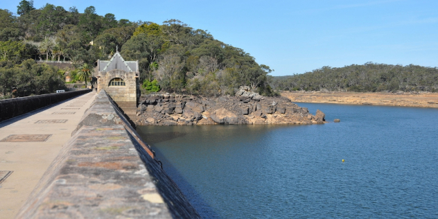 The view from a bridge along the top of a dam, with a stone gatehouse and tree-covered, rocky shore at the bridge's end.