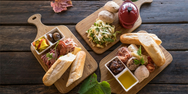 Overhead view of three light coloured wood charcuterie boards in a circle, holding cut baguette bread, pickles, oils and meats, on a dark wood table. 