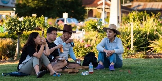 Two couples sit and drink out of cans on the grass with a bushy garden on a sunny day during the National Cherry Festival. 