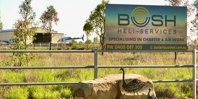 Bush heli-services sign on fence with emu and chick underneath and helicopter and hangar in background