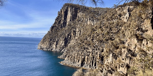 Craggy cliffs covered with patches of green and brown scrub rising up from dark blue water with no beach, against a blue sky. 