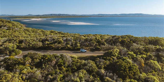  A panoramic view of a car driving along a forested road, with a large bay, and mountains in the distance beyond it.