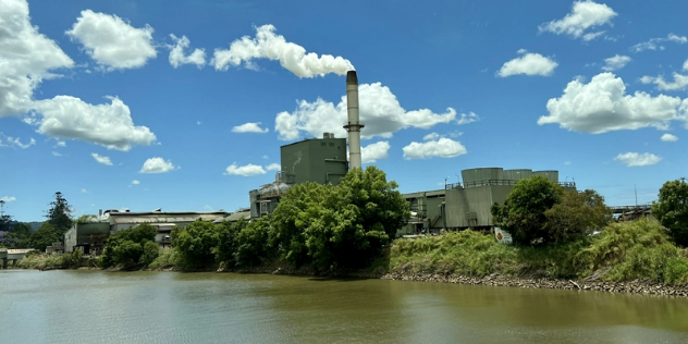  A metal industrial building with a tall silo built along a tree-lined riverbank, under blue skies. 