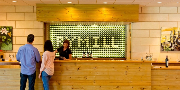 a man and a woman stand at a large pine bar while a woman in a black short sleeved top pours wine