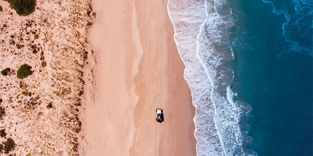 arial view of a white four-wheel drive vehicle driving along the beach with the ocean to the right