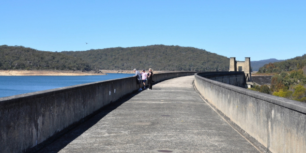  Three people at the concrete railing of a long curving bridge over the top of a dam, surrounded by green hills and blue sky. 