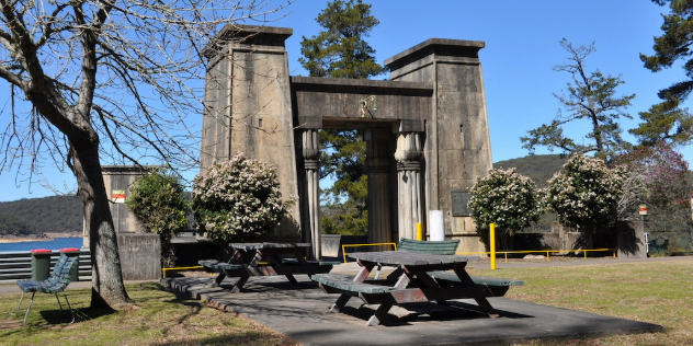  Two pillars with decorative columns forming a gate to a dam, with a sunny park and picnic tables in front of it. 