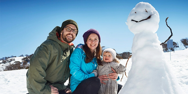  A man and woman holding a baby crouched next to a smiling snowman on a sunny, snow-covered hill. 