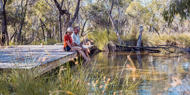  A middle-aged couple sit on a wooden pier, dipping their feet into a pool surrounded by trees.