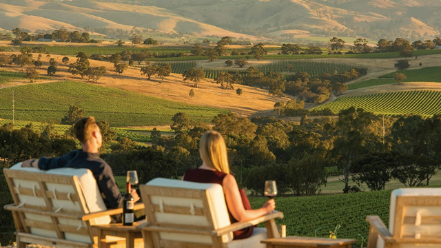couple-at-winery-barossa-sa-640x360 A man and a woman sit in chairs on a hill drinking wine at sunset, overlooking green hills and vineyards.